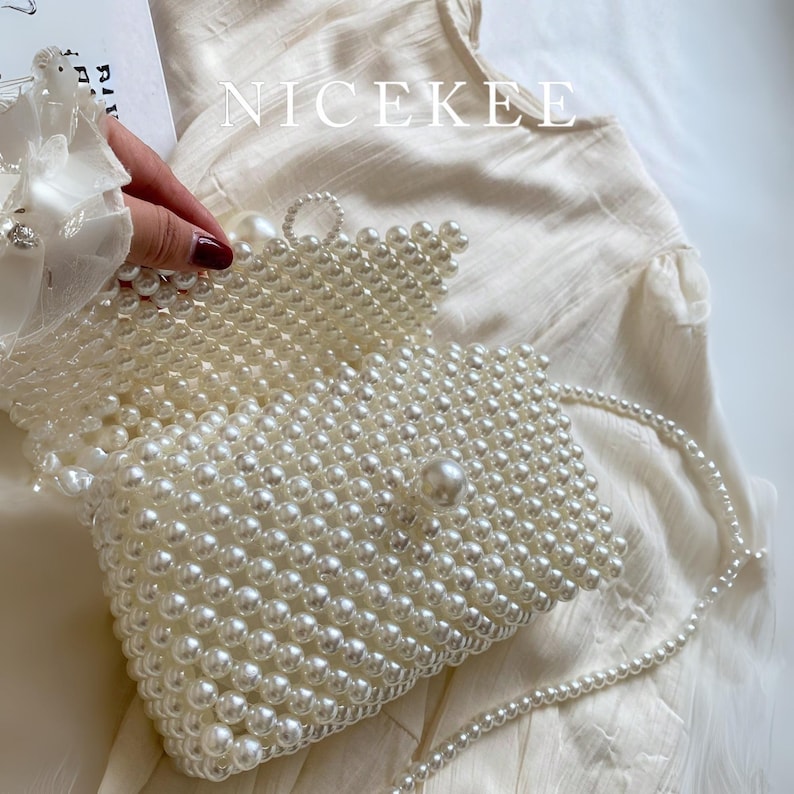 May include: A white beaded clutch bag with a pearl strap. The rectangular bag is covered in small, round, white beads. The flap is secured with a large pearl. The word "NICEKEE" is printed in white on the top of the image.