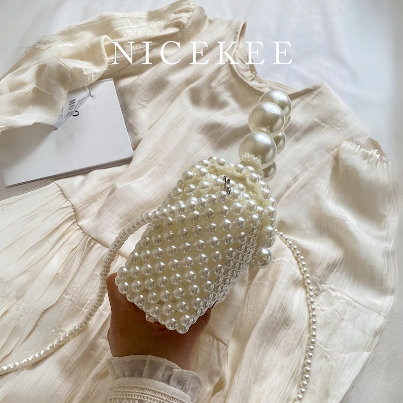 May include: A white beaded handbag with a pearl handle and a long pearl strap. The bag is cylindrical and covered in small, round beads. The word "NICEKEE" is printed in white across the top of the image.