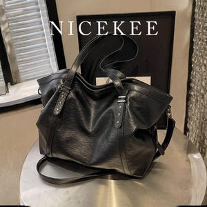 Puede incluir: Bolso tote de cuero negro con asa superior y bandolera desmontable. El bolso presenta herrajes plateados, incluyendo tachuelas en forma de estrella en las correas. La palabra "NICEKEE" está impresa en blanco en la parte superior.