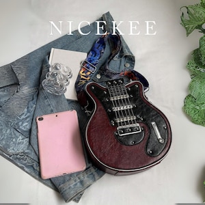 May include: A burgundy and black guitar-shaped handbag with a patterned strap rests on a denim jacket. A pink tablet, a small white book, and a clear glass object are also present. The word "NICEKEE" is at the top.