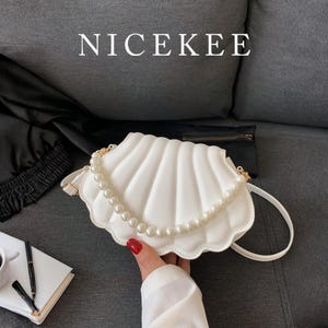 May include: A white seashell-shaped handbag with a pearl strap and gold-tone hardware. The bag is held by a person with red nail polish. The word "NICEKEE" is printed at the top. A cup of coffee and a notebook are in the background.