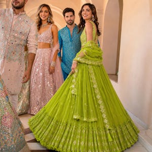 May include: A lime green lehenga set featuring a sleeveless blouse and a voluminous pleated skirt with intricate embroidery and mirror work. The ensemble includes a matching sheer dupatta draped over the shoulders, showcasing detailed borders and embellishments.