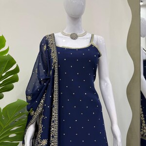 Pu&ograve; includere: Un abito blu navy con uno scialle trasparente abbinato &egrave; esposto su un manichino. L'abito presenta uno scollo quadrato con finiture dorate e piccoli abbellimenti dorati. Lo scialle e l'abito hanno dettagli ricamati in oro.