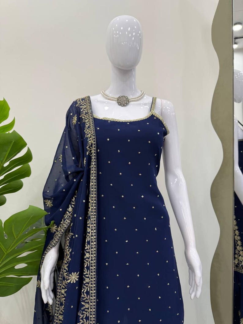 Pu&ograve; includere: Un abito blu navy con uno scialle trasparente abbinato &egrave; esposto su un manichino. L'abito presenta uno scollo quadrato con finiture dorate e piccoli abbellimenti dorati. Lo scialle e l'abito hanno dettagli ricamati in oro.