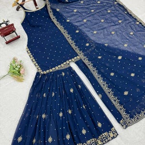 Pu&ograve; includere: Un completo etnico blu navy con top senza maniche e pantaloni svasati, ornato da ricami dorati e piccoli dettagli. Una sciarpa trasparente abbinata con dettagli simili completa l'ensemble. L'outfit &egrave; esposto su una gruccia di legno.
