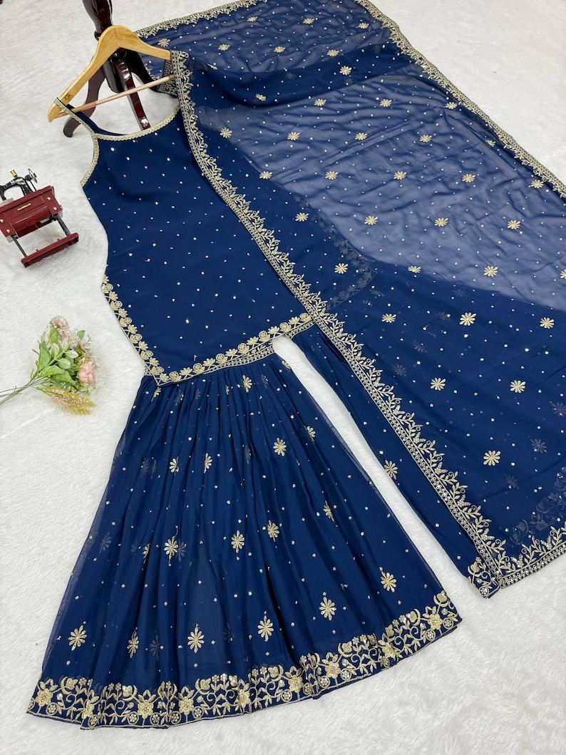 Pu&ograve; includere: Un completo etnico blu navy con top senza maniche e pantaloni svasati, ornato da ricami dorati e piccoli dettagli. Una sciarpa trasparente abbinata con dettagli simili completa l'ensemble. L'outfit &egrave; esposto su una gruccia di legno.