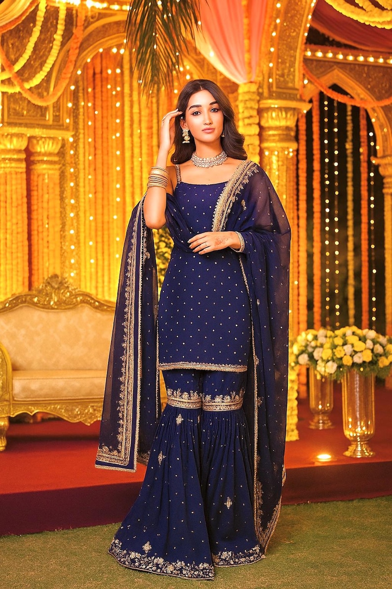 Pu&ograve; includere: Una persona indossa un salwar kameez blu navy con ricami dorati. L'outfit comprende un top, pantaloni svasati e un dupatta trasparente. Lo sfondo presenta decorazioni dorate e composizioni floreali, per una festa.
