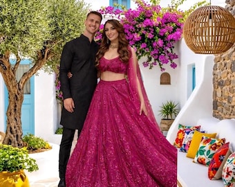 May include: A couple poses outdoors. The woman wears a magenta-colored, embellished skirt and crop top with a matching sheer scarf. The man wears a black tunic and pants. The background includes a white building, bougainvillea, and a decorative hanging light.
