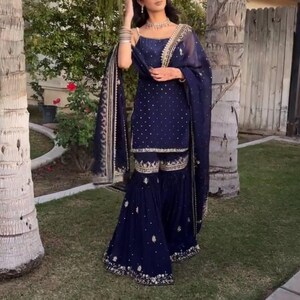 Pu&ograve; includere: Un completo salwar kameez blu navy. L'outfit comprende un top, pantaloni svasati e una sciarpa abbinata. Gli indumenti sono adornati da intricati ricami dorati e piccoli abbellimenti bianchi sparsi. L'outfit &egrave; indossato con gioielli.