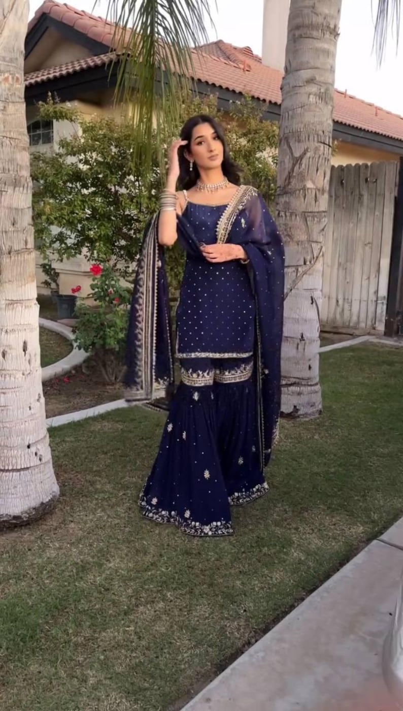 Pu&ograve; includere: Un completo salwar kameez blu navy. L'outfit comprende un top, pantaloni svasati e una sciarpa abbinata. Gli indumenti sono adornati da intricati ricami dorati e piccoli abbellimenti bianchi sparsi. L'outfit &egrave; indossato con gioielli.