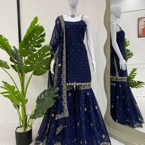 Pu&ograve; includere: Un completo Sharara blu navy esposto su un manichino. Il completo presenta un top senza maniche e pantaloni svasati, entrambi adornati con ricami dorati e piccoli punti dorati sparsi. Una dupatta abbinata &egrave; drappeggiata sulla spalla. L'outfit &egrave; esposto davanti a uno specchio.