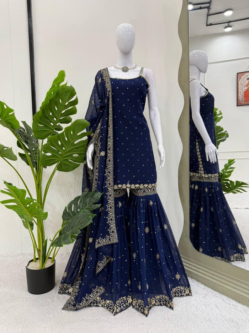 Pu&ograve; includere: Un completo Sharara blu navy esposto su un manichino. Il completo presenta un top senza maniche e pantaloni svasati, entrambi adornati con ricami dorati e piccoli punti dorati sparsi. Una dupatta abbinata &egrave; drappeggiata sulla spalla. L'outfit &egrave; esposto davanti a uno specchio.