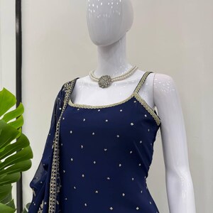 Pu&ograve; includere: Un completo blu navy con un top a collo quadrato e uno scialle drappeggiato abbinato, ornato di dettagli dorati. L'outfit &egrave; impreziosito da piccoli pois color oro. Una collana di perle con un elemento centrale color argento completa il look.