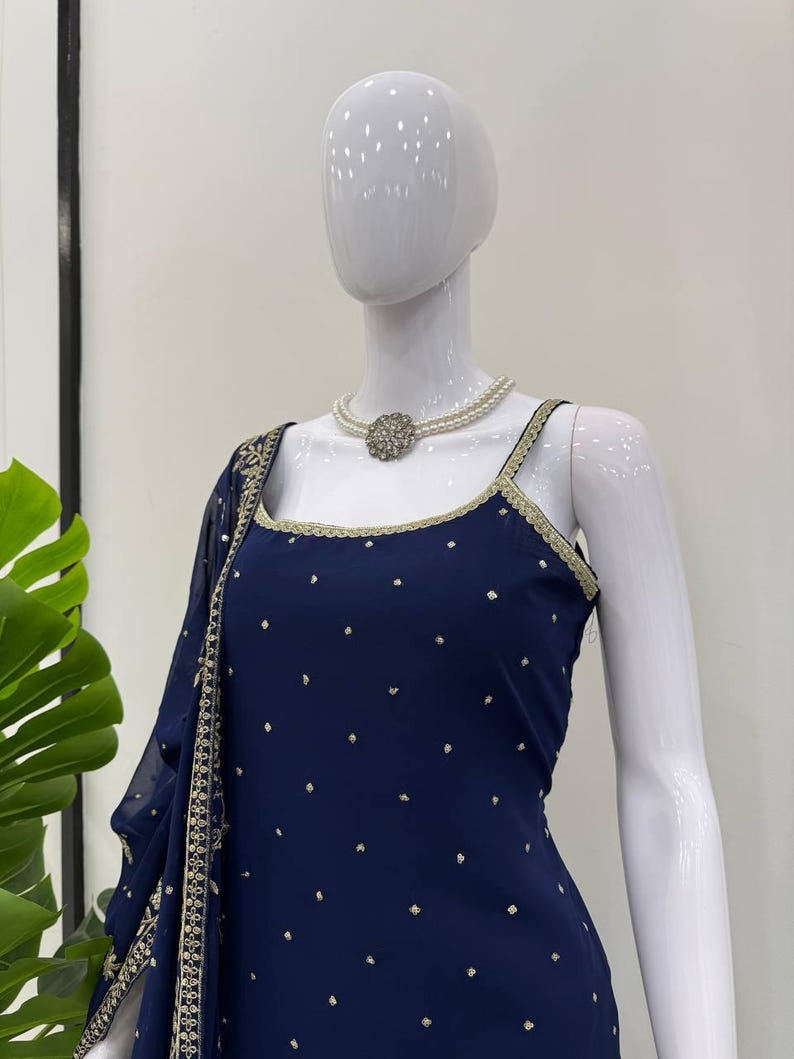Pu&ograve; includere: Un completo blu navy con un top a collo quadrato e uno scialle drappeggiato abbinato, ornato di dettagli dorati. L'outfit &egrave; impreziosito da piccoli pois color oro. Una collana di perle con un elemento centrale color argento completa il look.
