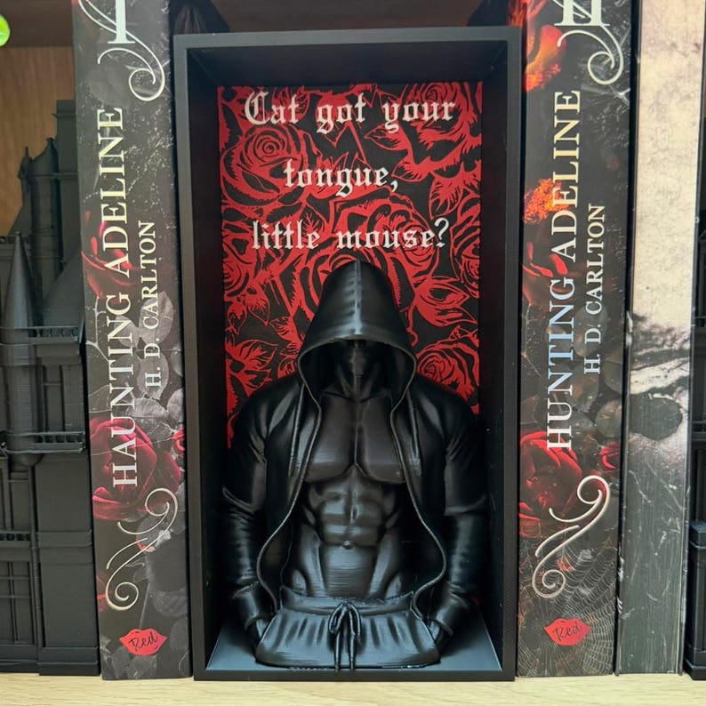 Puede incluir: Una escultura negra de una figura encapuchada con un torso musculoso, exhibida en una estanter&iacute;a negra. El fondo presenta un patr&oacute;n floral rojo con la cita "Cat got your tongue, little mouse?". A ambos lados de la estanter&iacute;a hay dos libros titulados "Haunting Adeline".