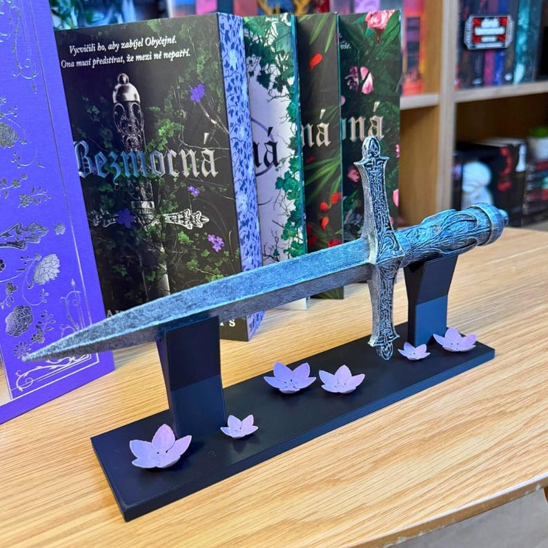 May include: A decorative sword with a detailed hilt and blade rests on a black stand adorned with purple flower accents. The sword is displayed in front of a row of books with floral designs and text.