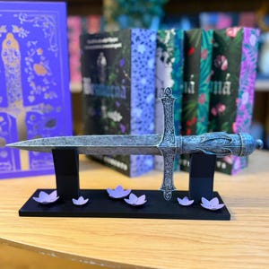 May include: A decorative silver dagger with an ornate handle and blade, displayed on a black stand with purple flower accents. The dagger is in front of a row of books with colorful covers, creating a fantasy theme.
