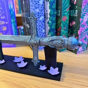 May include: A decorative sword bookend with a detailed, ornate design in shades of silver and blue. The bookend features a sword shape with a crossguard and handle. Small purple flower accents are placed on the black base. Books with colorful covers are in the background.
