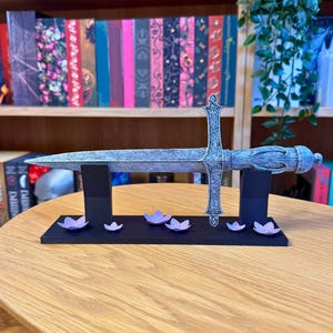 May include: A decorative silver sword with an ornate handle and crossguard, displayed on a black stand. The stand is decorated with small purple flower accents. The sword is resting on a wooden table.