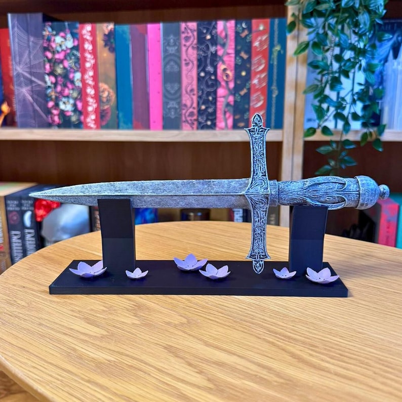 May include: A decorative silver sword with an ornate handle and crossguard, displayed on a black stand. The stand is decorated with small purple flower accents. The sword is resting on a wooden table.