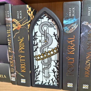 May include: A black, arched bookmark featuring a design of a silver snake entwined around a sword, a gold crown, and a white background. The bookmark is placed between the pages of a book with the title "KRUT&Yacute; PRINC".
