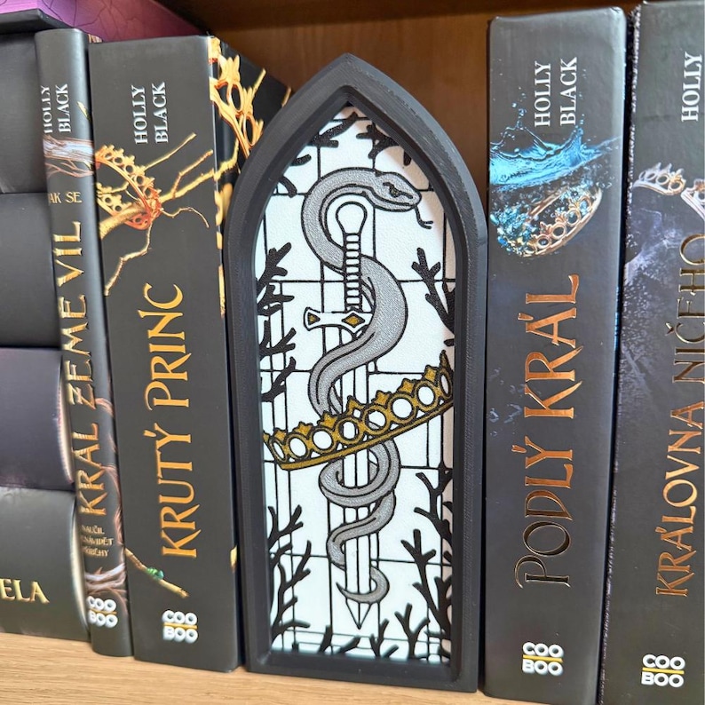 May include: A black, arched bookmark featuring a design of a silver snake entwined around a sword, a gold crown, and a white background. The bookmark is placed between the pages of a book with the title "KRUT&Yacute; PRINC".