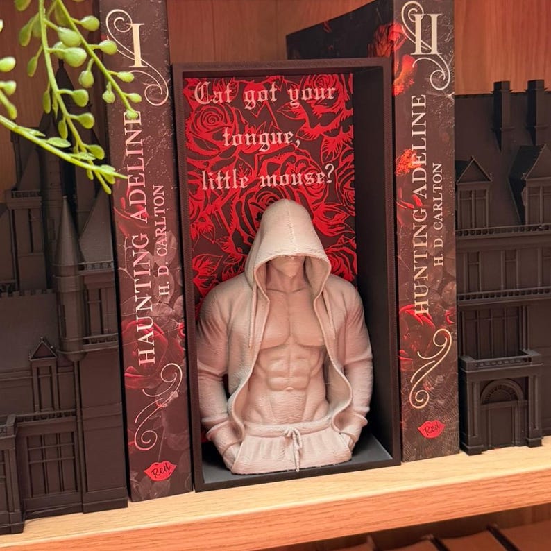 Puede incluir: Una escultura beige de una figura musculosa con sudadera con capucha y pantalones de ch&aacute;ndal, colocada entre dos libros titulados "Haunting Adeline". El fondo presenta un patr&oacute;n floral rojo con el texto "Cat got your tongue, little mouse?"