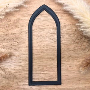May include: A dark navy blue arched window frame, likely for crafting or decoration, is centered on a light wood grain surface. The frame has a simple, elegant design. Dried ornamental grasses are in the background.