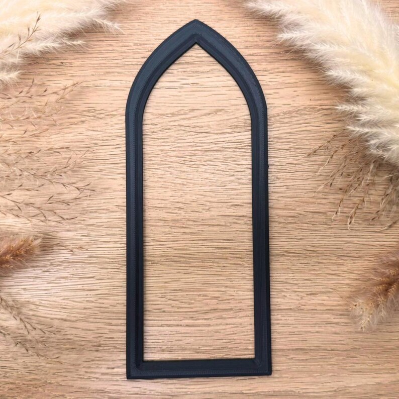 May include: A dark navy blue arched window frame, likely for crafting or decoration, is centered on a light wood grain surface. The frame has a simple, elegant design. Dried ornamental grasses are in the background.