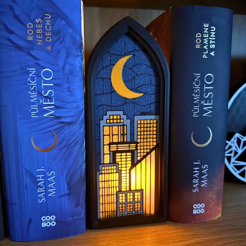 May include: A black, arched-shaped night light with a cityscape design. The night light features a crescent moon and city buildings with illuminated windows. The night light is placed between two books with the title "Půlměs&iacute;čn&iacute; Město" by Sarah J. Maas.