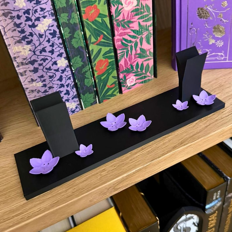 May include: A black wooden bookshelf display with two upright supports and several purple flower accents. The display is on a wooden shelf with books in the background. The books have colorful floral and botanical designs.