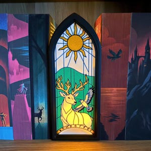 May include: A decorative stained glass-style bookend featuring a yellow deer, a bird, and a sun design. The bookend is framed in black and is illuminated. The bookend is placed between several books.
