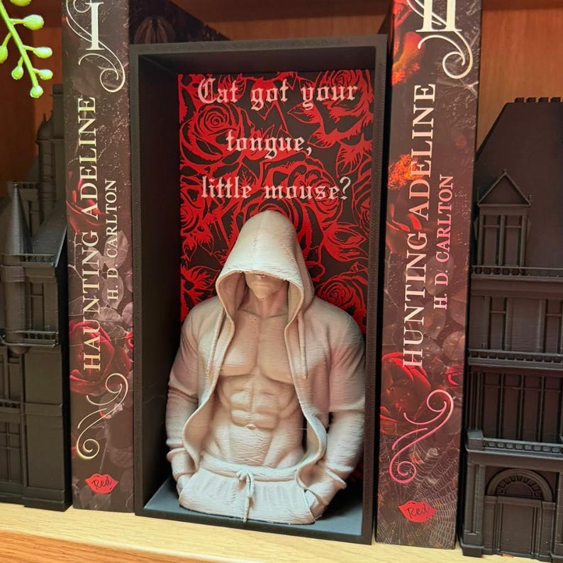 Puede incluir: Una escultura beige de una figura musculosa con una sudadera con capucha y pantalones de ch&aacute;ndal, exhibida en una estanter&iacute;a negra. La estanter&iacute;a est&aacute; flanqueada por dos libros titulados "HAUNTING ADELINE" y un tel&oacute;n de fondo con el texto "Cat got your tongue, little mouse?"
