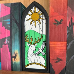 May include: A black framed bookmark with a stained glass design. The design features a golden sun, green hills, a white deer with golden antlers, and a white dove. The bookmark is placed between the pages of a book.
