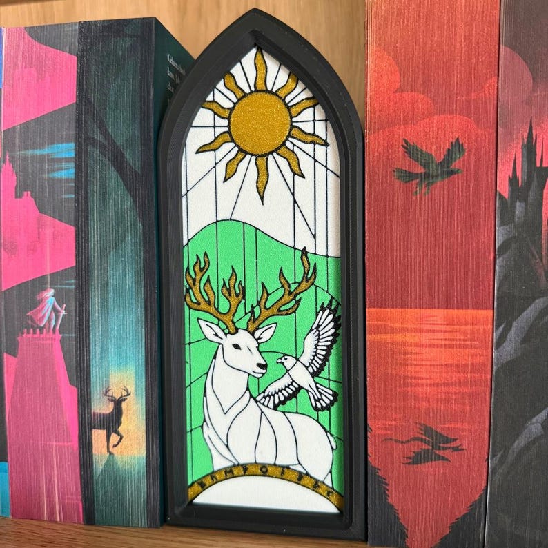 May include: A black framed bookmark with a stained glass design. The design features a golden sun, green hills, a white deer with golden antlers, and a white dove. The bookmark is placed between the pages of a book.