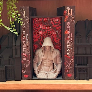 Puede incluir: Una escultura beige de una persona con sudadera con capucha y pantal&oacute;n de ch&aacute;ndal, con torso musculoso, se exhibe entre dos sujetalibros. Los sujetalibros est&aacute;n dise&ntilde;ados como libros con el t&iacute;tulo "HAUNTING ADELINE" y el texto "Cat got your tongue, little mouse?"