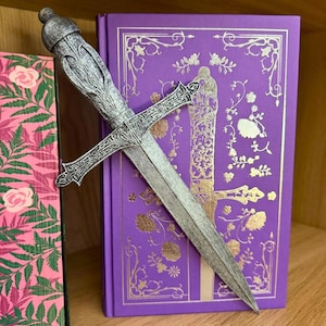 May include: A decorative silver letter opener in the shape of a sword rests on a purple book with gold floral accents. The letter opener has an ornate handle and a long, pointed blade. The book is next to a pink book with a floral pattern.