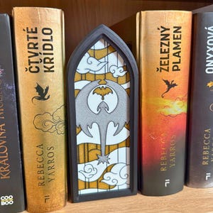 May include: A decorative bookmark with a stained-glass design featuring a gray dragon silhouette against a yellow and white background. The bookmark is framed in black and is placed between books with gold and orange spines.