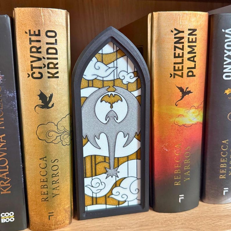 May include: A decorative bookmark with a stained-glass design featuring a gray dragon silhouette against a yellow and white background. The bookmark is framed in black and is placed between books with gold and orange spines.