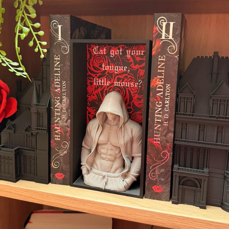 Puede incluir: Una estanter&iacute;a decorativa con una figura musculosa con capucha, flanqueada por dos libros titulados "HAUNTING ADELINE" y un letrero con el texto "Cat got your tongue, little mouse?". Los libros y la exhibici&oacute;n son oscuros.