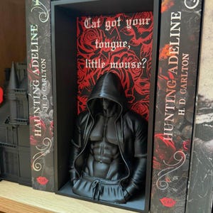Puede incluir: Una escultura negra de una figura encapuchada con un torso musculoso, colocada entre dos libros titulados "HAUNTING ADELINE". El fondo presenta un patr&oacute;n floral rojo y negro con el texto "Cat got your tongue, little mouse?"