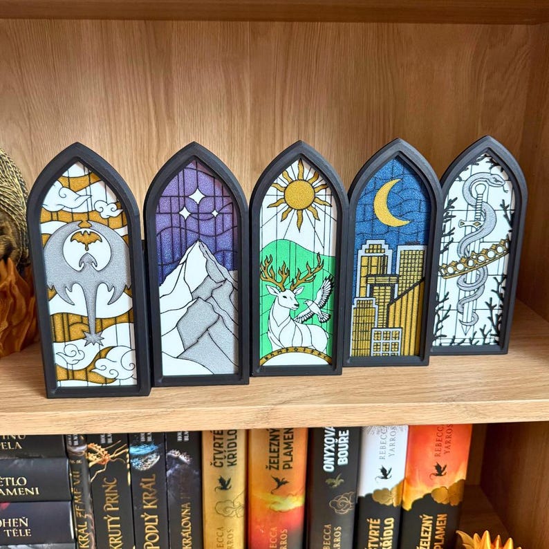 May include: Five decorative stained glass panels with black frames, each featuring a unique design. The panels depict a variety of scenes, including a mountain, a deer, a cityscape, and celestial elements. They are displayed on a wooden shelf.