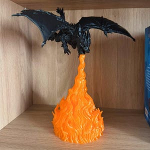 May include: A black dragon figurine with outstretched wings, perched above a vibrant orange flame. The dragon is detailed with textured scales and a menacing pose. The flame is sculpted to appear as if it is erupting from the dragon's mouth. The figurine is displayed on a wooden shelf.