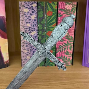 May include: A decorative, silver-colored sword with an ornate handle and crossguard is positioned in front of a row of books. The books have colorful floral and patterned covers in shades of purple, green, and pink. The sword's design is detailed.
