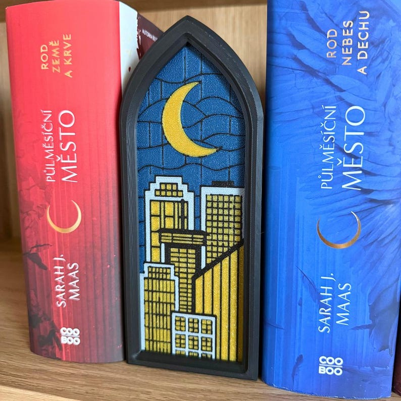 May include: A black bookmark with a stained glass design featuring a yellow crescent moon over a cityscape. The bookmark is between two books with the text "PŮLMĚS&Iacute;ČN&Iacute; MĚSTO" and "SARAH J. MAAS" on the spines.