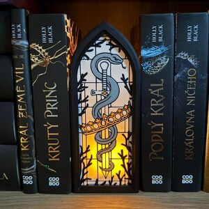 May include: A black, arched-shaped decorative light with a stained-glass design featuring a snake coiled around a sword, a crown, and a yellow glow. The light is placed in front of a row of black books with gold lettering.