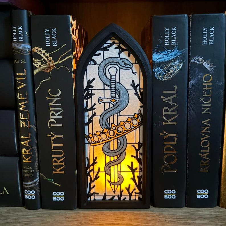 May include: A black, arched-shaped decorative light with a stained-glass design featuring a snake coiled around a sword, a crown, and a yellow glow. The light is placed in front of a row of black books with gold lettering.