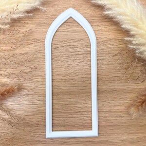 May include: A white, arched window-shaped cookie cutter. The cutter is made of plastic and has a rectangular shape with a pointed arch at the top. The cutter is on a wooden surface.