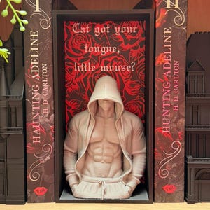 Puede incluir: Escultura beige de una figura musculosa con sudadera con capucha, colocada entre dos libros titulados "Haunting Adeline". El fondo presenta un dise&ntilde;o floral rojo y el texto "Cat got your tongue, little mouse?"
