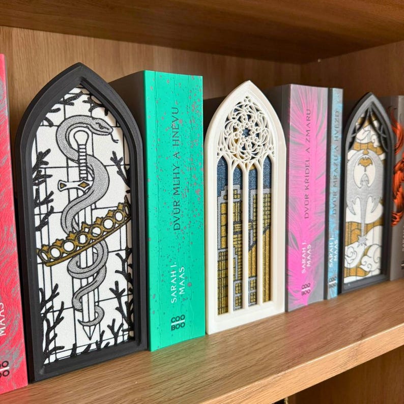 May include: A collection of decorative bookends in a row on a wooden shelf. The bookends feature intricate designs, including a sword and crown, a gothic window, and other ornate patterns. The books have titles in a foreign language.
